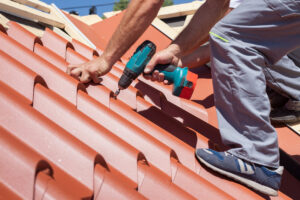 Worker on a roof with electric drill installing red metal tile on wooden house